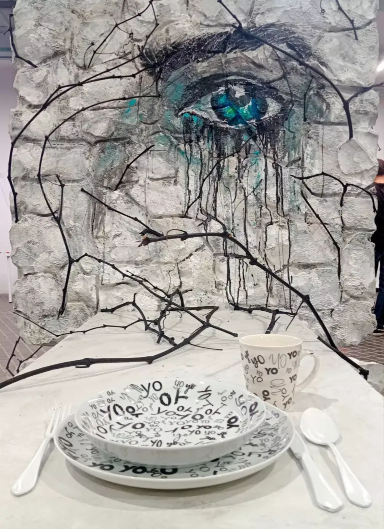 Pieza artística compuesta por una mesa con platos, vaso y cubiertos, y en frente una pared decorada con una ilustración de un ojo que te mira